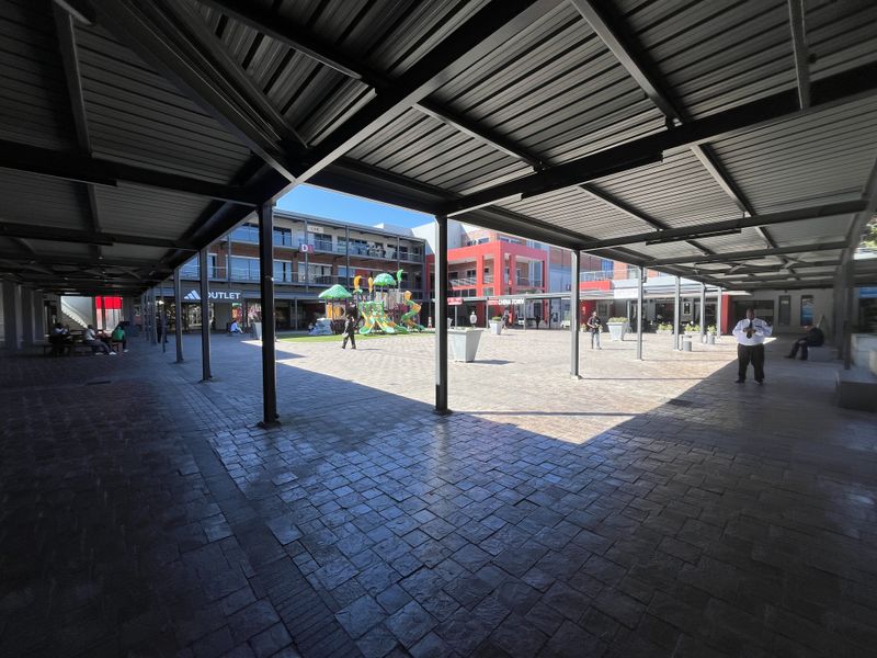 A spacious covered outdoor retail plaza featuring paved flooring and a modern steel roof structure, providing a sheltered walkway between commercial storefronts.