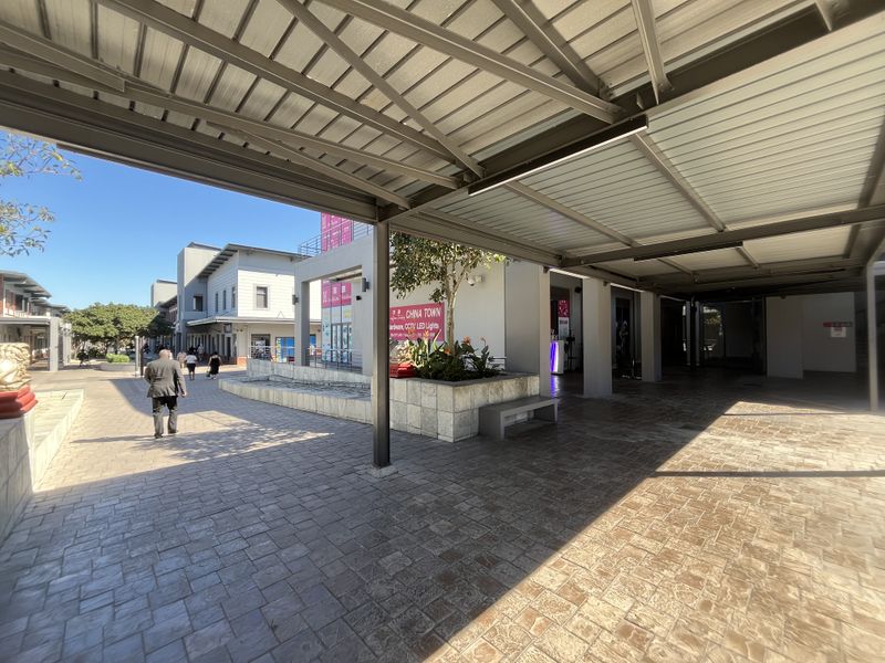 A well-maintained outdoor retail plaza featuring a covered walkway, paved pedestrian areas, and integrated landscaping with built-in seating.
