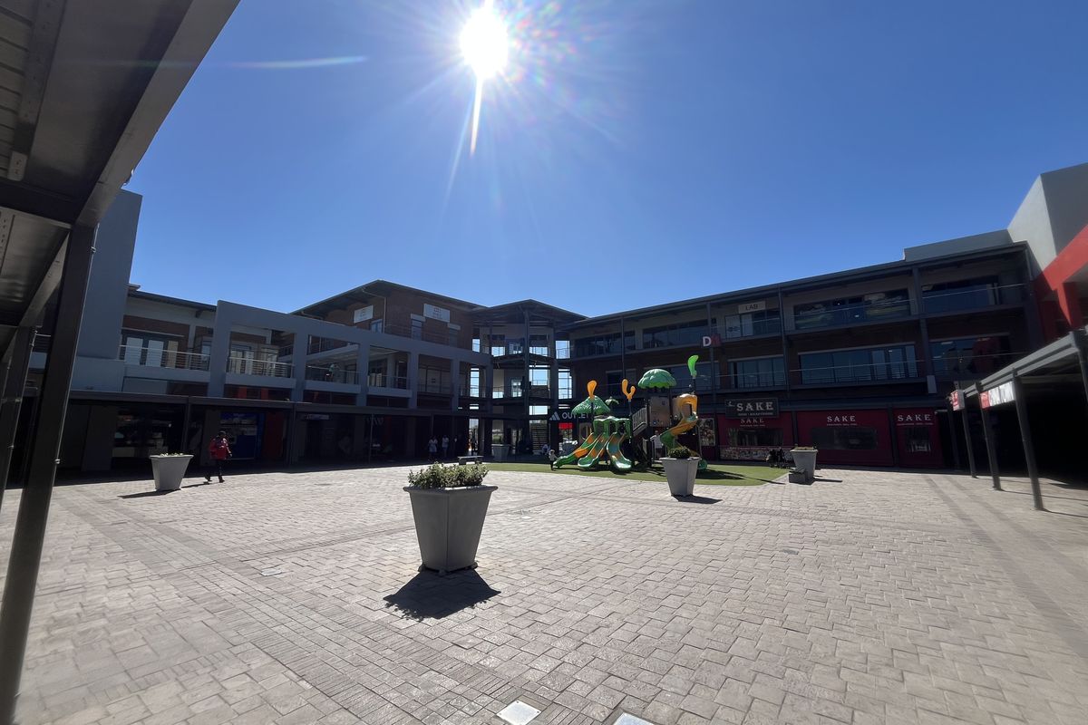 A wide-angle view of a modern retail center courtyard featuring paved walkways, multi-level commercial units, and structural steel architectural elements.