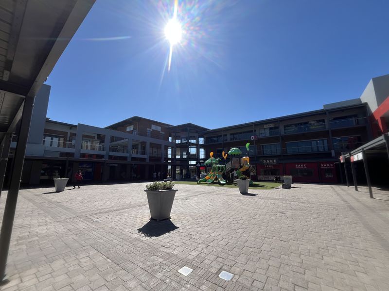 A wide-angle view of a modern retail center courtyard featuring paved walkways, multi-level commercial units, and structural steel architectural elements.