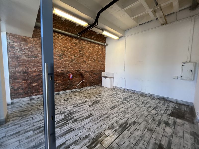 Open-plan retail space featuring an exposed brick wall, wood-look tile flooring, industrial-style exposed ceiling, and a small kitchenette area.