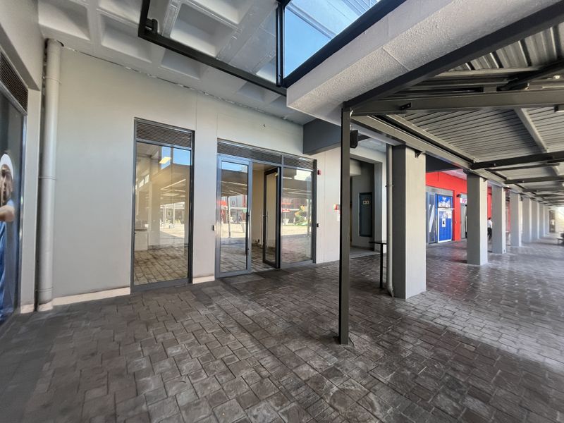 A covered outdoor walkway area featuring paved flooring, glass storefront entrances, and a modern metal roof structure with integrated skylights.
