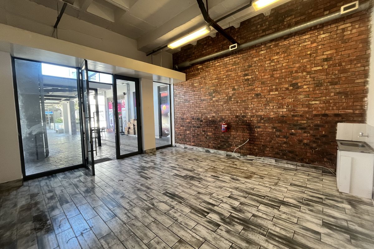 Open-plan retail space featuring exposed brick walls, industrial concrete ceilings, tiled flooring, and a glass storefront entrance with a small kitchenette area.