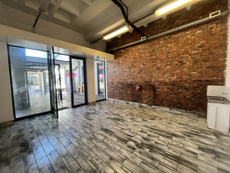 Open-plan retail space featuring exposed brick walls, industrial concrete ceilings, tiled flooring, and a glass storefront entrance with a small kitchenette area.