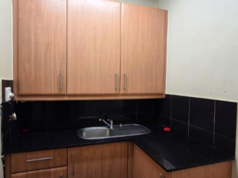 A compact office kitchenette featuring wood-look laminate cabinetry, a black granite countertop, and a stainless steel sink with a black tiled backsplash.