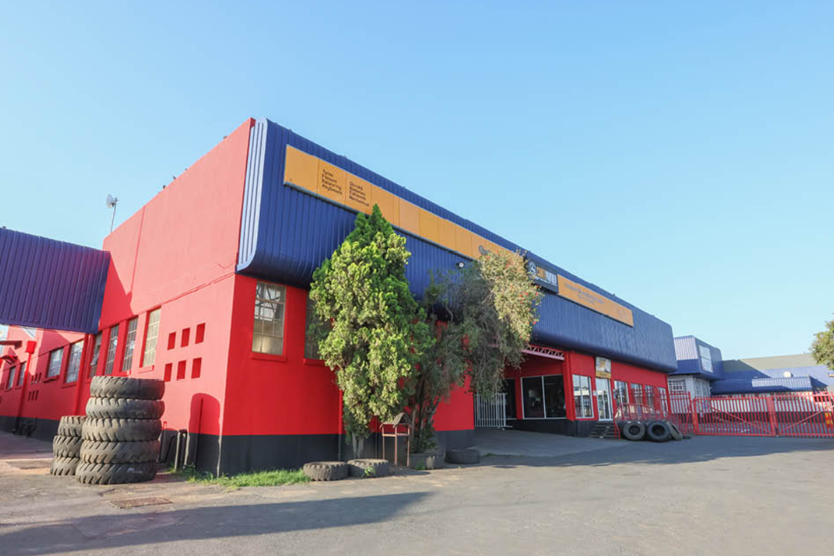 Front exterior view of a commercial industrial property featuring prominent red and blue metal cladding, asphalt driveway, and secure perimeter fencing.