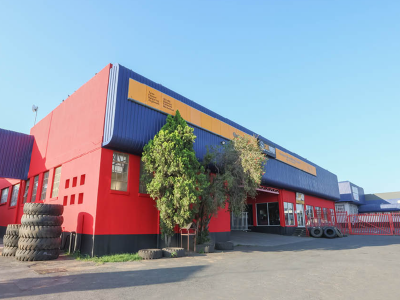Front exterior view of a commercial industrial property featuring prominent red and blue metal cladding, asphalt driveway, and secure perimeter fencing.