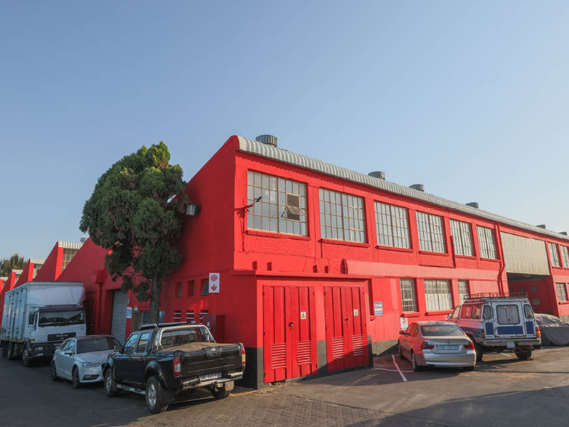 Exterior view of a large industrial warehouse facility featuring prominent red masonry walls, steel-framed windows, and multiple roller shutter access points.