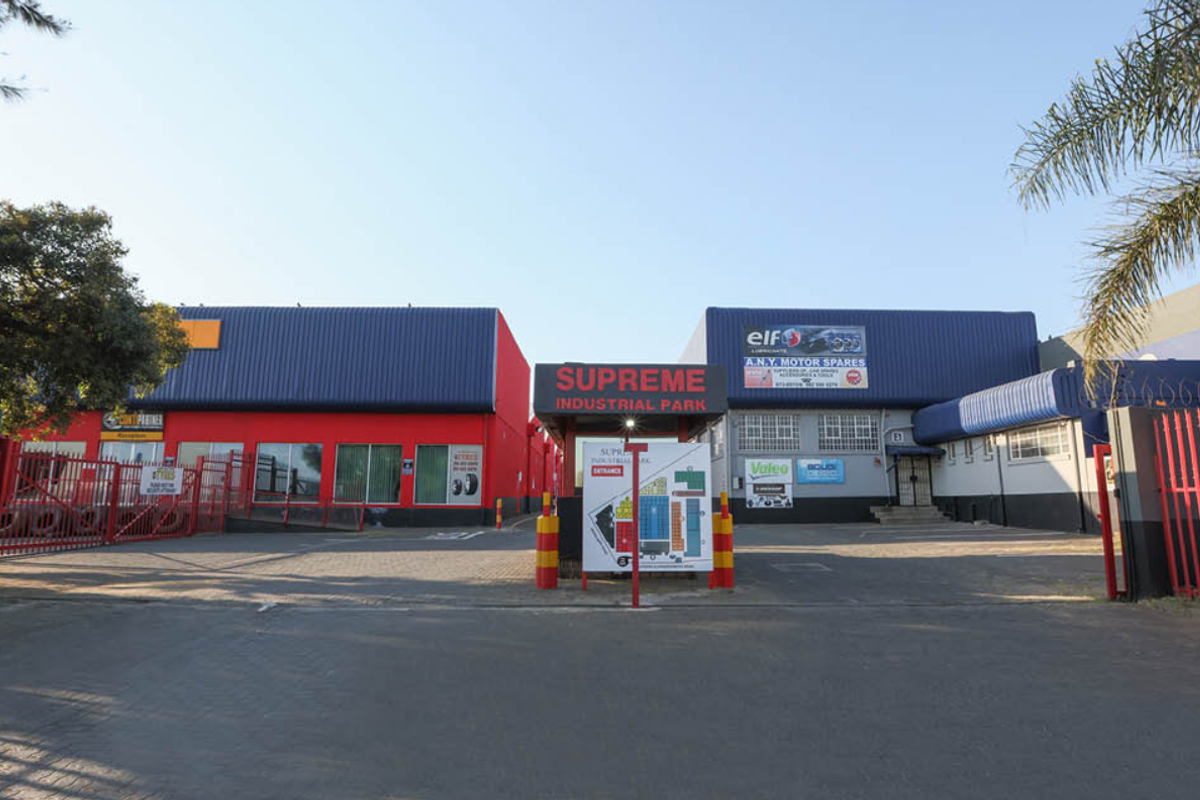 Front entrance view of Supreme Industrial Park featuring secure gated access, asphalt driveway, and multiple industrial warehouse units with prominent signage.