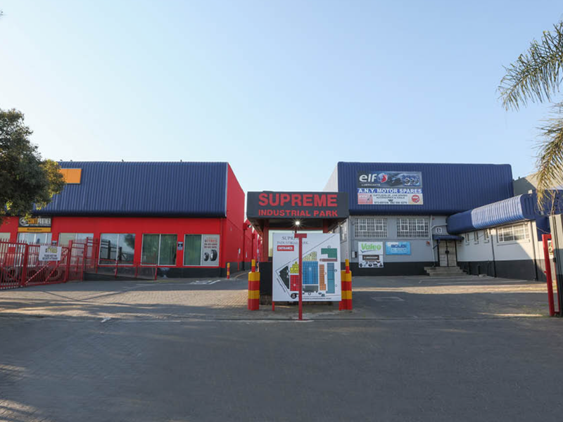 Front entrance view of Supreme Industrial Park featuring secure gated access, asphalt driveway, and multiple industrial warehouse units with prominent signage.