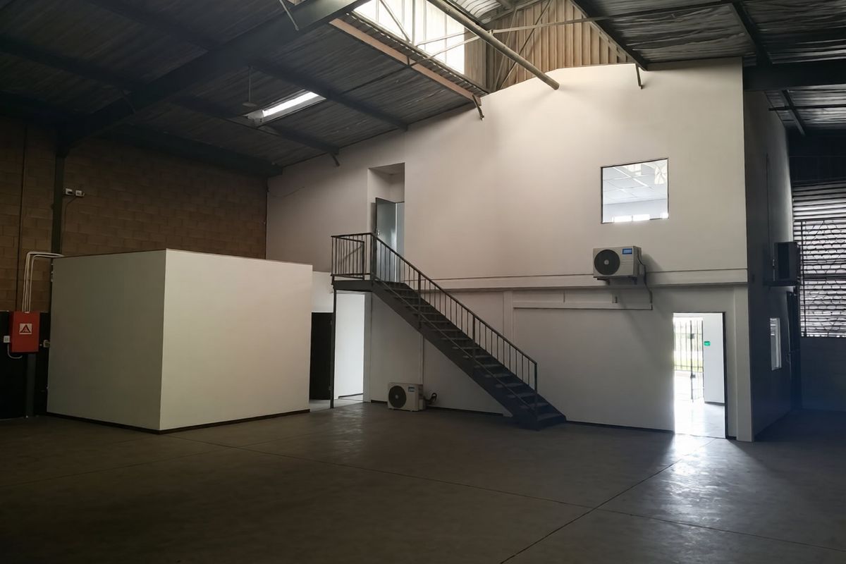 Spacious industrial warehouse unit featuring high ceilings with exposed steel trusses, concrete flooring, a mezzanine staircase, and integrated air conditioning units.