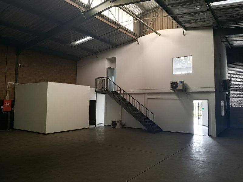 Spacious industrial warehouse unit featuring high ceilings with exposed steel trusses, concrete flooring, a mezzanine staircase, and integrated air conditioning units.