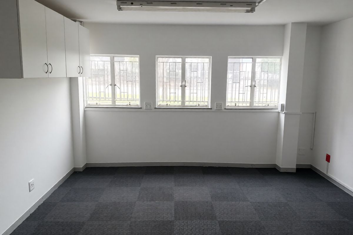 A clean, empty office space featuring carpet tiles, built-in wall cabinetry, and windows fitted with security bars.