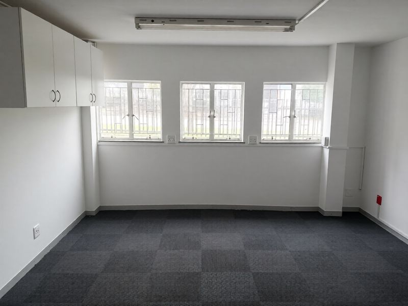 A clean, empty office space featuring carpet tiles, built-in wall cabinetry, and windows fitted with security bars.