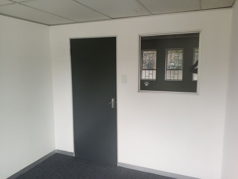 A clean, empty office space featuring neutral walls, carpet flooring, a suspended ceiling, and an internal window providing visibility into adjacent areas.