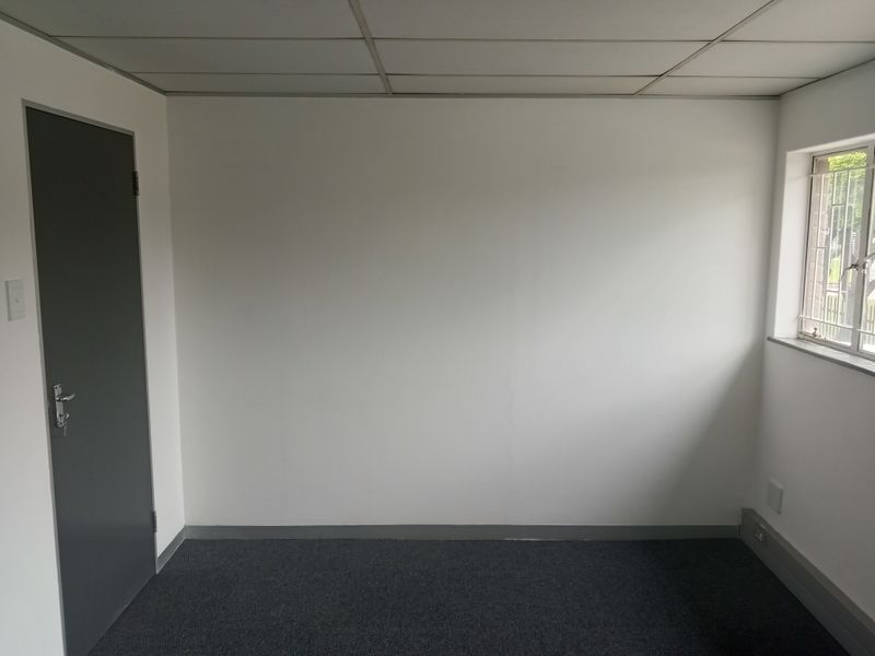 A clean, empty office space featuring neutral walls, carpeted flooring, a suspended ceiling, and a window with security bars.