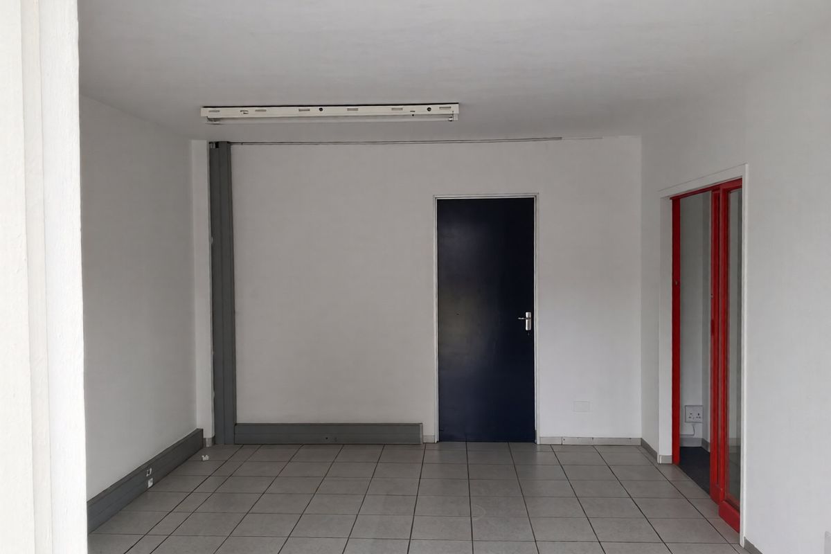 An empty, functional office space featuring tiled flooring, a fluorescent light fixture, and a red-framed glass partition.