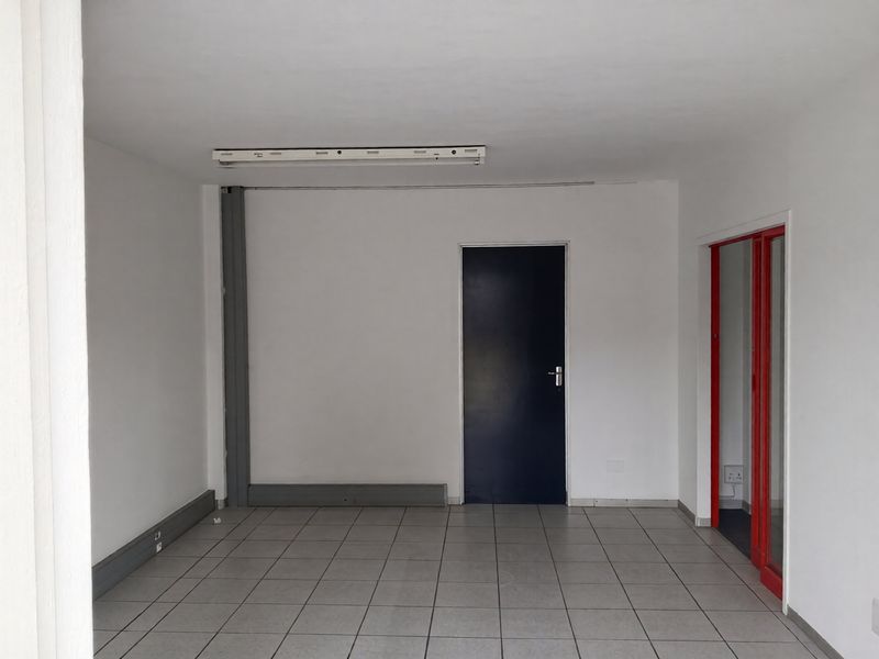 An empty, functional office space featuring tiled flooring, a fluorescent light fixture, and a red-framed glass partition.