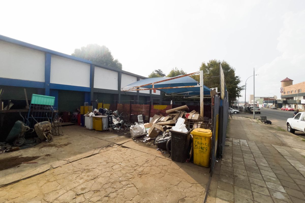 Exterior view of an industrial property featuring a concrete yard, metal shade structure, and roller shutter doors, showing significant signs of wear and debris.