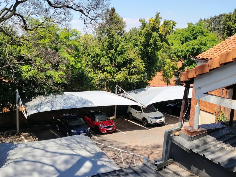 Elevated view of a paved parking area featuring multiple shade cloth carports, surrounded by mature trees and adjacent to the building structure.