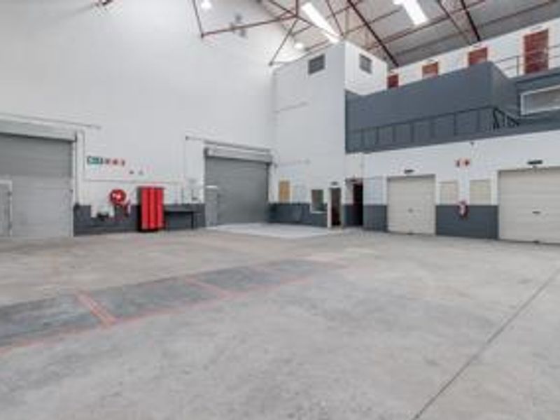 Spacious industrial warehouse unit featuring high ceilings with exposed trusses, multiple roller shutter doors, concrete flooring, and a mezzanine level for additional storage or office.