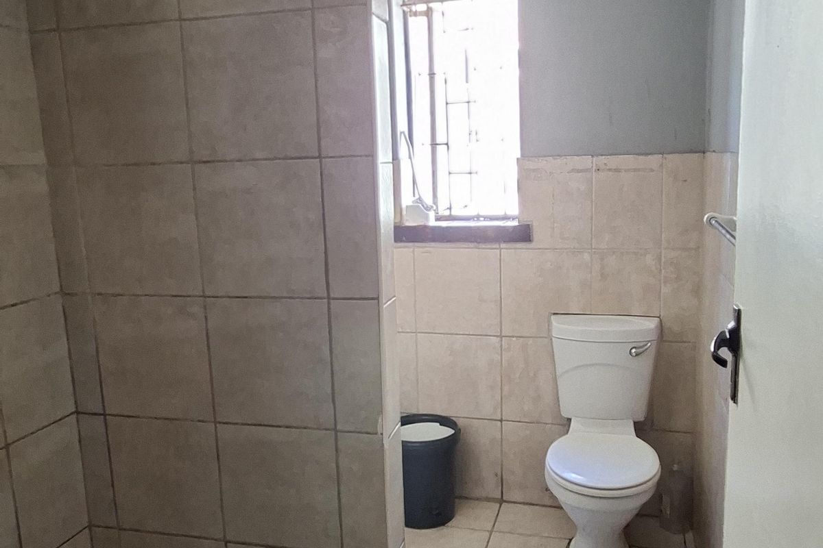 A functional bathroom featuring a tiled shower enclosure, toilet, and an exposed electric geyser, requiring cosmetic updates to improve its overall condition.