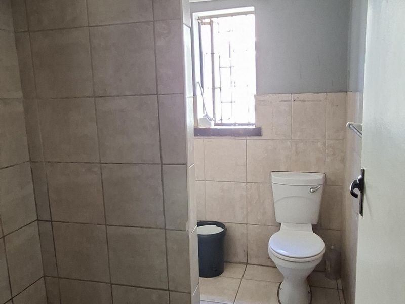 A functional bathroom featuring a tiled shower enclosure, toilet, and an exposed electric geyser, requiring cosmetic updates to improve its overall condition.