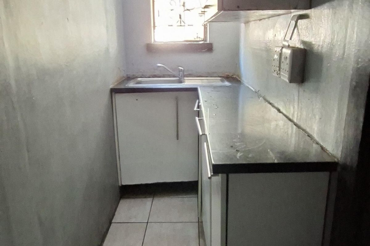 A compact, narrow kitchen featuring tiled flooring, built-in cabinetry with laminate countertops, and a single stainless steel sink under a window.