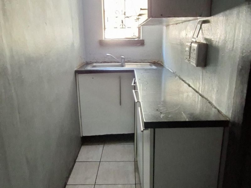 A compact, narrow kitchen featuring tiled flooring, built-in cabinetry with laminate countertops, and a single stainless steel sink under a window.