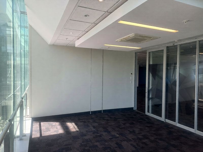 A modern, vacant office space featuring floor-to-ceiling glass walls, carpeted flooring, suspended ceilings with recessed lighting, and integrated air conditioning.
