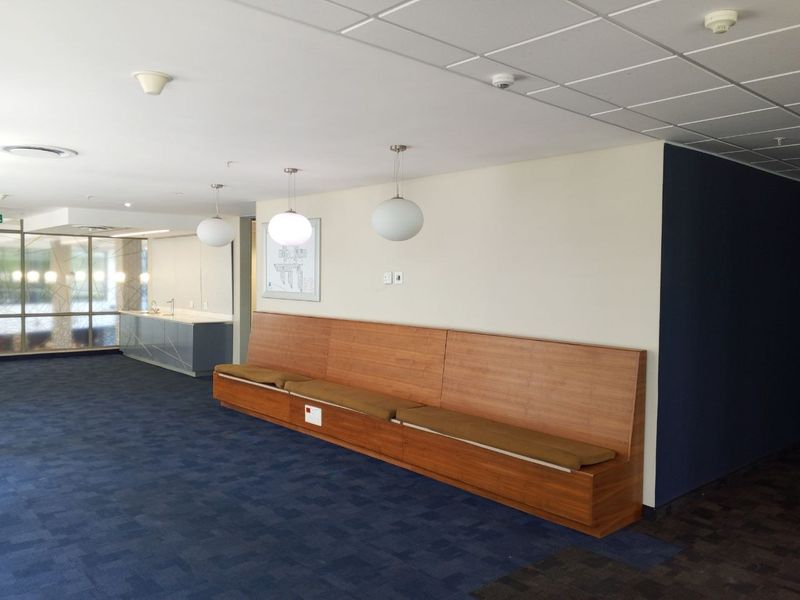 Modern office reception area featuring built-in wooden bench seating, blue carpet tiles, drop ceiling, and a small kitchenette area with a sink.