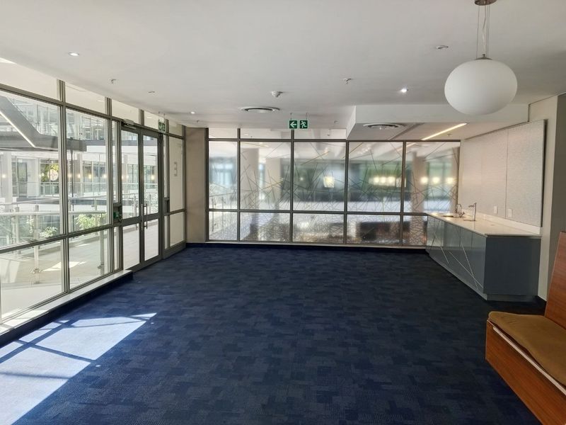 A bright, open-plan office space featuring large glass partitions, blue carpeting, and a built-in kitchenette with a sink.