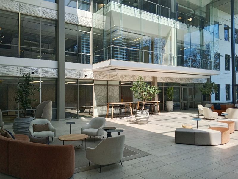 A modern, light-filled open-plan office atrium featuring expansive glass walls, high ceilings, and contemporary tiled flooring in a professional corporate environment.