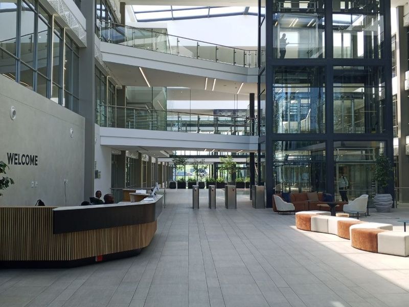 Modern office building atrium featuring a glass elevator, multi-level walkways, high ceilings with skylights, and a professional reception area.