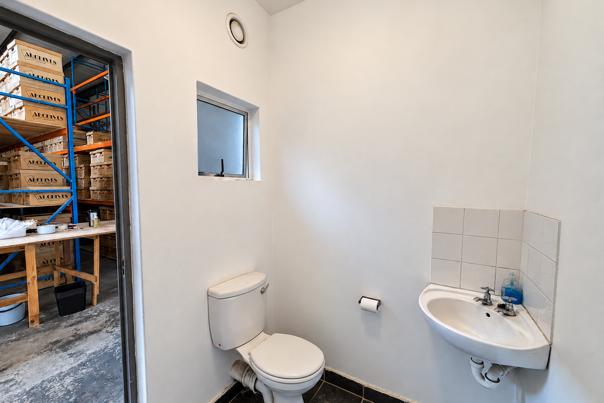 A clean, functional industrial-style restroom featuring a wall-mounted basin with tiled splashback and a standard toilet, adjacent to a warehouse storage area.