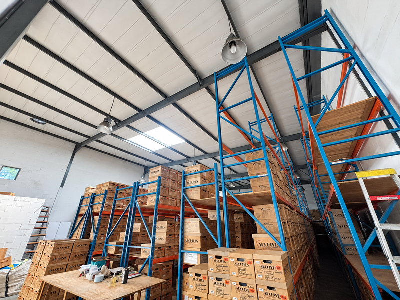 Industrial warehouse space featuring high ceilings with exposed steel trusses, skylights, and extensive built-in steel racking systems for storage.