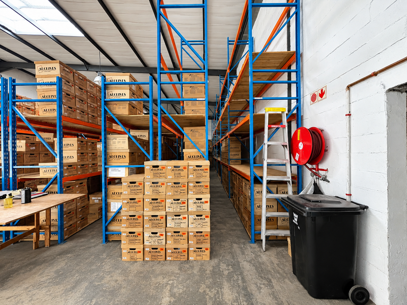 Industrial warehouse space featuring extensive steel racking systems, exposed roof trusses with skylights, and a wall-mounted fire hose reel.