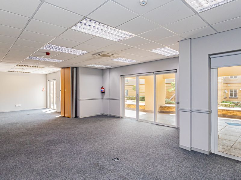 A spacious, empty office area featuring a suspended ceiling with recessed lighting, carpeted flooring, and large sliding glass doors providing access to an outdoor area.