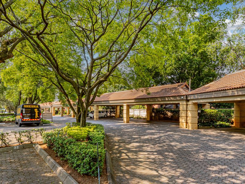 A well-maintained office exterior featuring covered parking bays with brick pillars, a paved driveway, and mature trees in a landscaped setting.