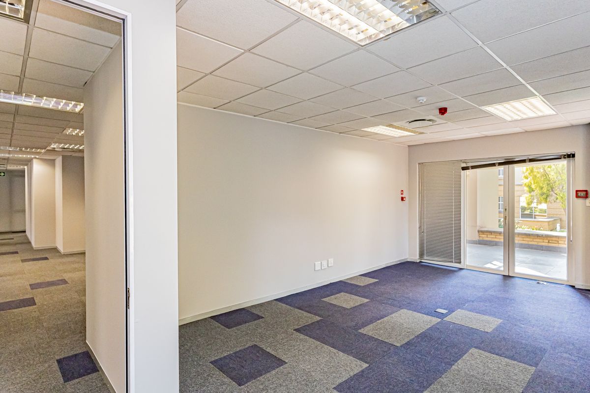 A bright, empty office space featuring carpet tiles, a suspended ceiling with recessed lighting, and a sliding glass door leading to an outdoor area.