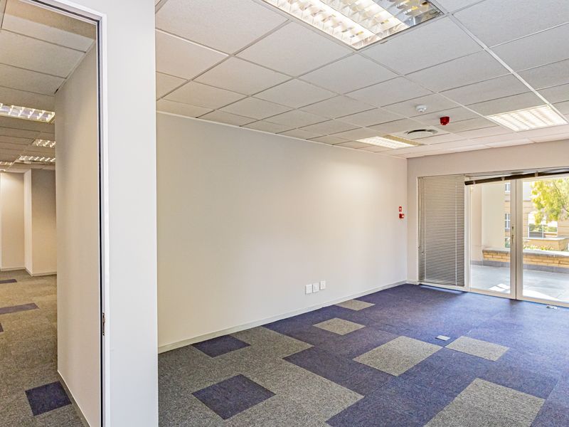A bright, empty office space featuring carpet tiles, a suspended ceiling with recessed lighting, and a sliding glass door leading to an outdoor area.