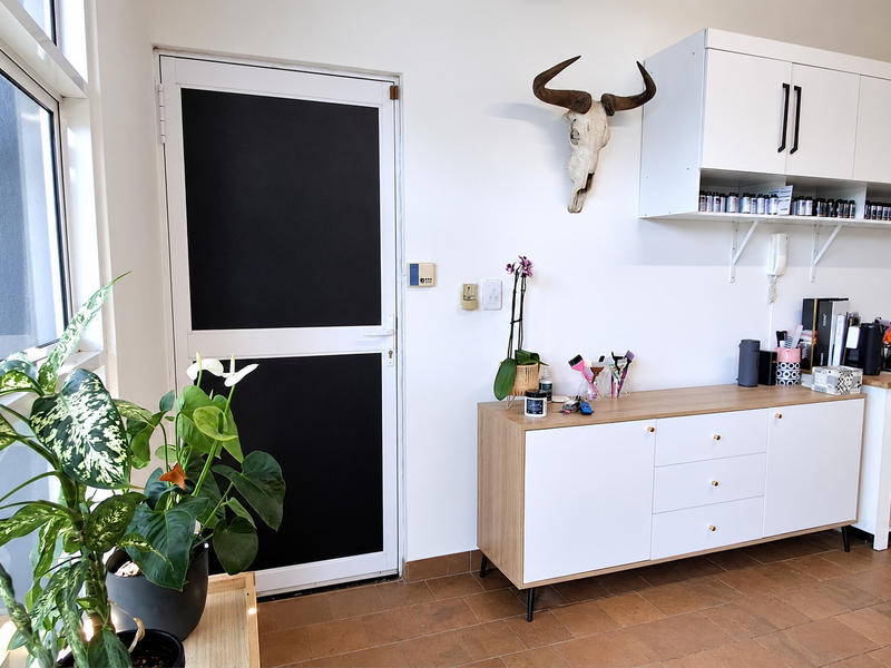 A bright office space featuring built-in wall cabinetry, tiled flooring, and a modern aluminium framed door with natural light from a large window.