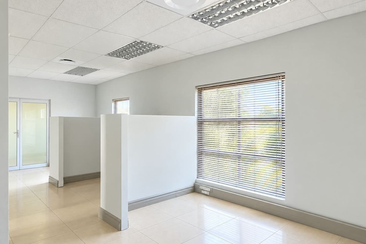 A bright, open-plan office space featuring tiled flooring, suspended ceilings with recessed lighting, partition walls, and large windows with fitted venetian blinds.