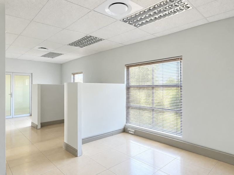 A bright, open-plan office space featuring tiled flooring, suspended ceilings with recessed lighting, partition walls, and large windows with fitted venetian blinds.