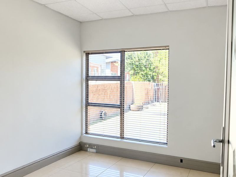 A bright, empty office space featuring tiled flooring, a suspended ceiling with recessed lighting, and a large window with blinds.