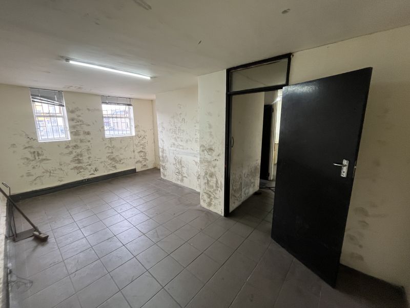 An empty office space featuring tiled flooring, fluorescent lighting, and security bars on windows, currently requiring significant wall repairs and cosmetic refurbishment.