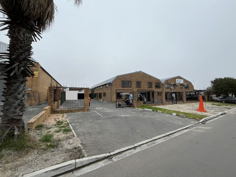Front exterior view of an industrial property featuring brick construction, secure gated access, paved driveway, and wall-mounted air conditioning units.