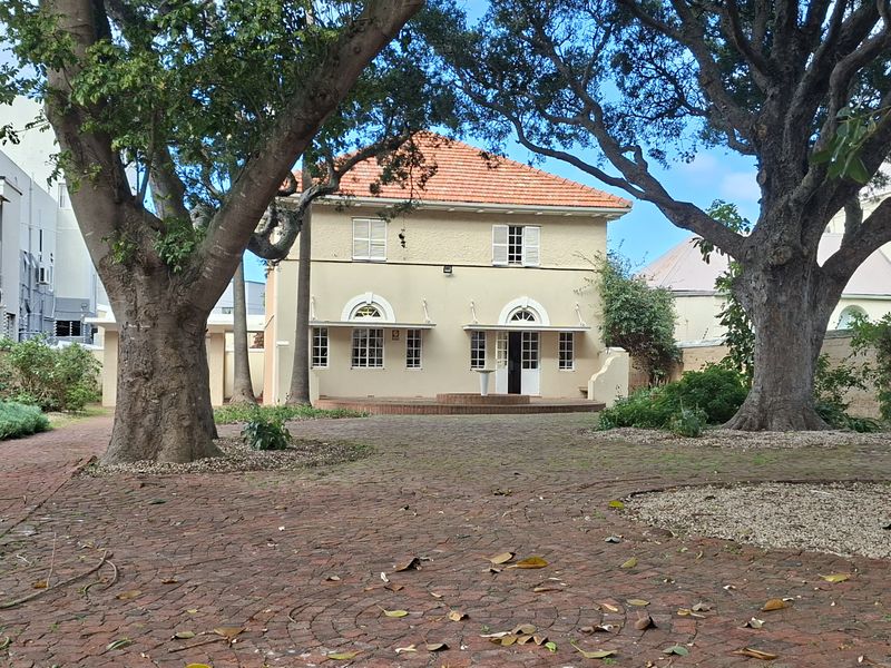 A rear view of the office property featuring a spacious brick-paved courtyard, mature trees, and a classic building facade with terracotta roof tiles.