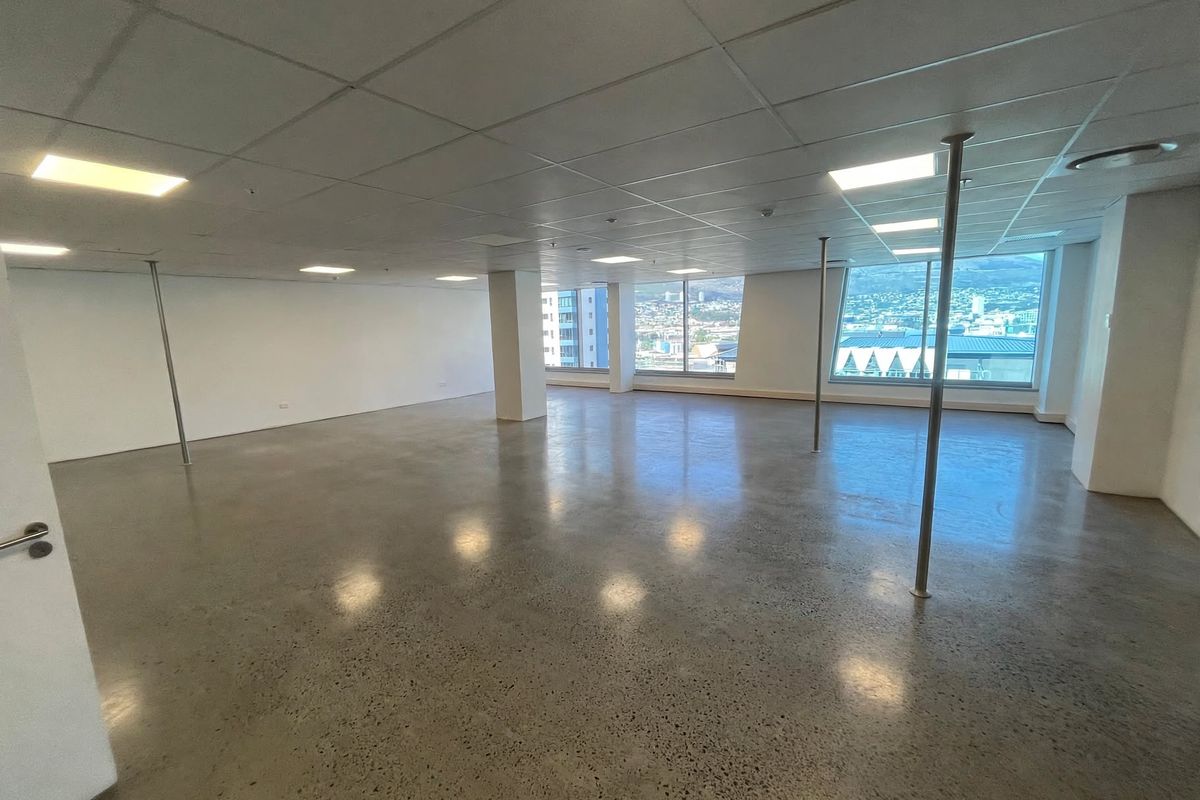 A spacious, open-plan office area featuring polished concrete floors, suspended ceiling tiles, recessed lighting, and large windows offering expansive city views.