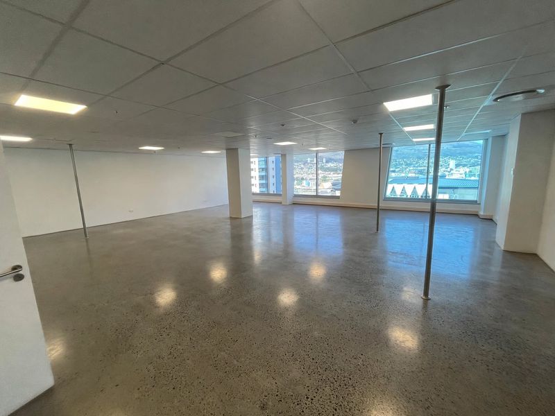 A spacious, open-plan office area featuring polished concrete floors, suspended ceiling tiles, recessed lighting, and large windows offering expansive city views.